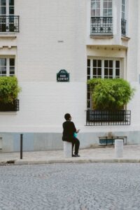 Vue arrière d'une personne assise sur une borne de rue sur la rue Cortot, cadre urbain avec bâtiment blanc.
