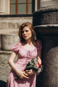Young woman in a pink dress leaning on stone pillar holding a bouquet, outdoors in Türkiye.