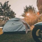 Tente de camping blanche près d'un lac, soleil couchant, moto à l'avant-plan