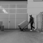 Homme pousse chariot avec carton devant entrepôt, clôture et cartons empilés à proximité