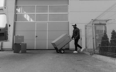 Homme pousse chariot avec carton devant entrepôt, clôture et cartons empilés à proximité