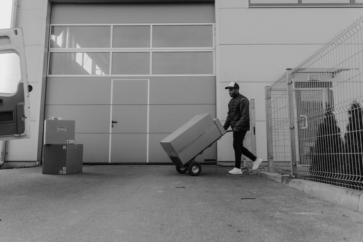 Homme pousse chariot avec carton devant entrepôt, clôture et cartons empilés à proximité