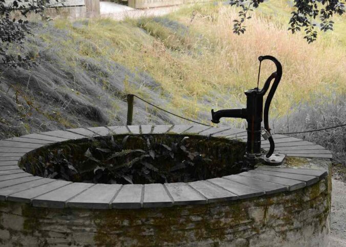 Un puits en pierre avec pompe manuelle, eau et colline herbeuse